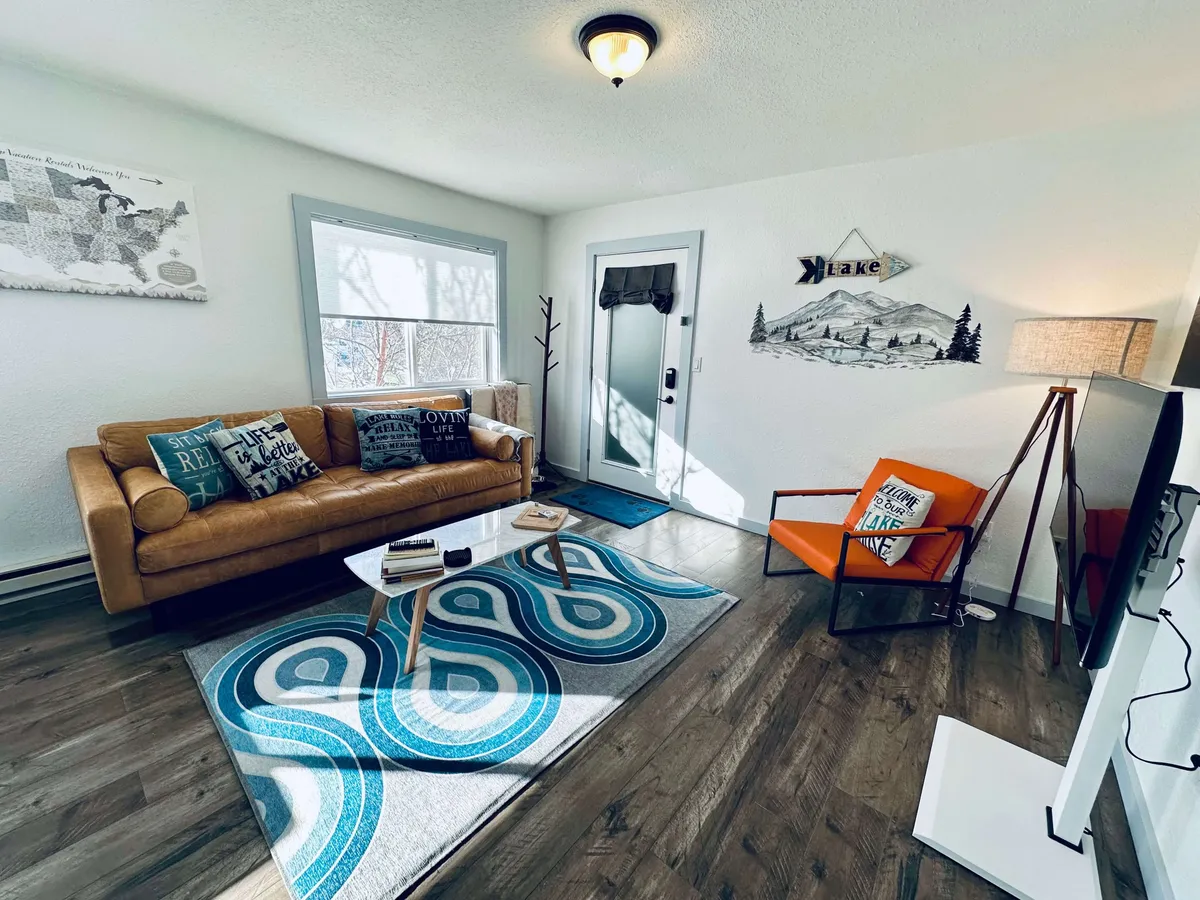 Another angle of the living area with a plush sofa, a striking orange chair, a central coffee table, patterned area rug, and vibrant wall art. The space is styled for both relaxation and group gatherings, with natural light enhancing its warmth.