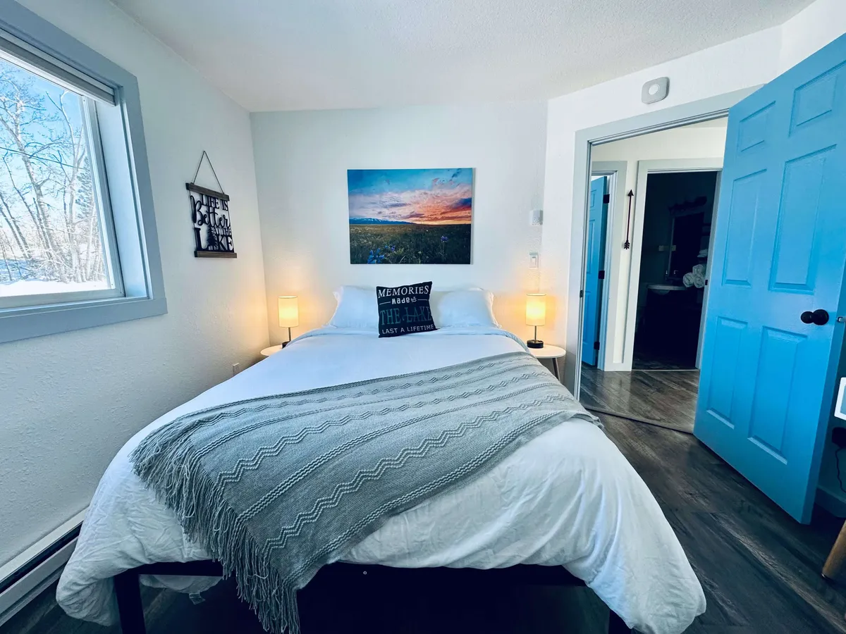 This image shows a cozy bedroom with a comfortable bed, soft lighting from lamps, and tasteful artwork on the wall. Natural light streams in through a window, creating a peaceful atmosphere, while the blue door adds a modern touch.