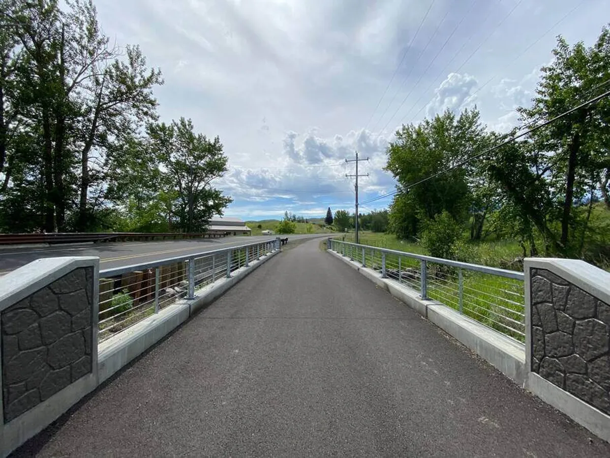A peaceful bridge surrounded by leafy trees under a bright blue sky, with walkers and cyclists enjoying the pathway. The scene captures the gateway to local trails and bike paths, providing easy access to Joseph’s natural wonders and fostering a spirit of exploration.