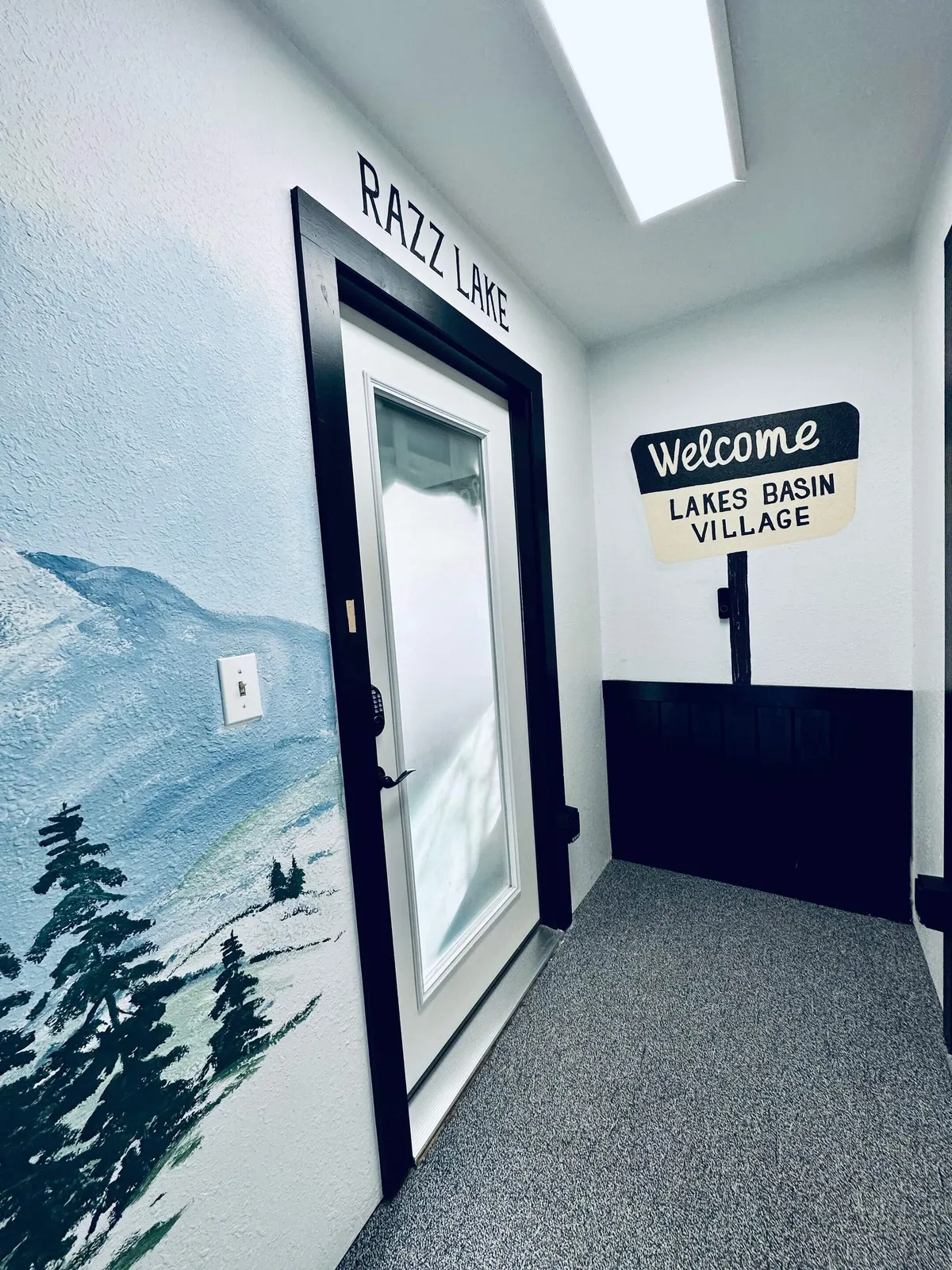 The entryway area welcoming adventurers, adorned with a mural of mountains and trees, a door labeled 'RAZZ LAKE,' and a sign greeting guests to Lakes Basin Village. The setting hints at outdoor exploration and serves as a transition from adventure to comfort.