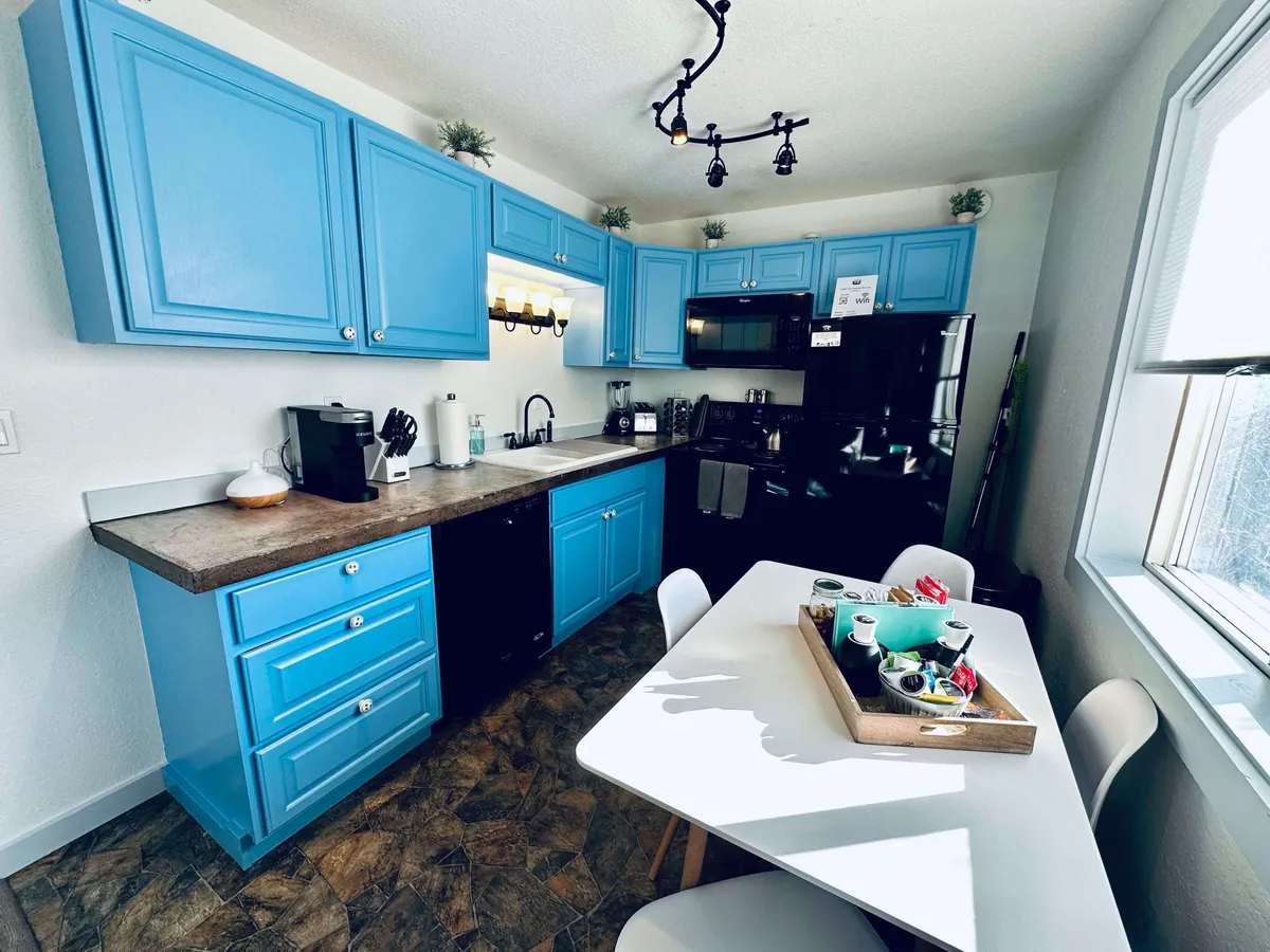 A spacious, cheerful kitchen is the focal point, featuring bright blue cabinets, sleek black appliances, and a dining table set with a tray of items. The open layout suggests both functionality and warmth, perfect for group cooking or shared meals. The scene is lively, evoking the bustle and joy of meal prep and communal dining.
