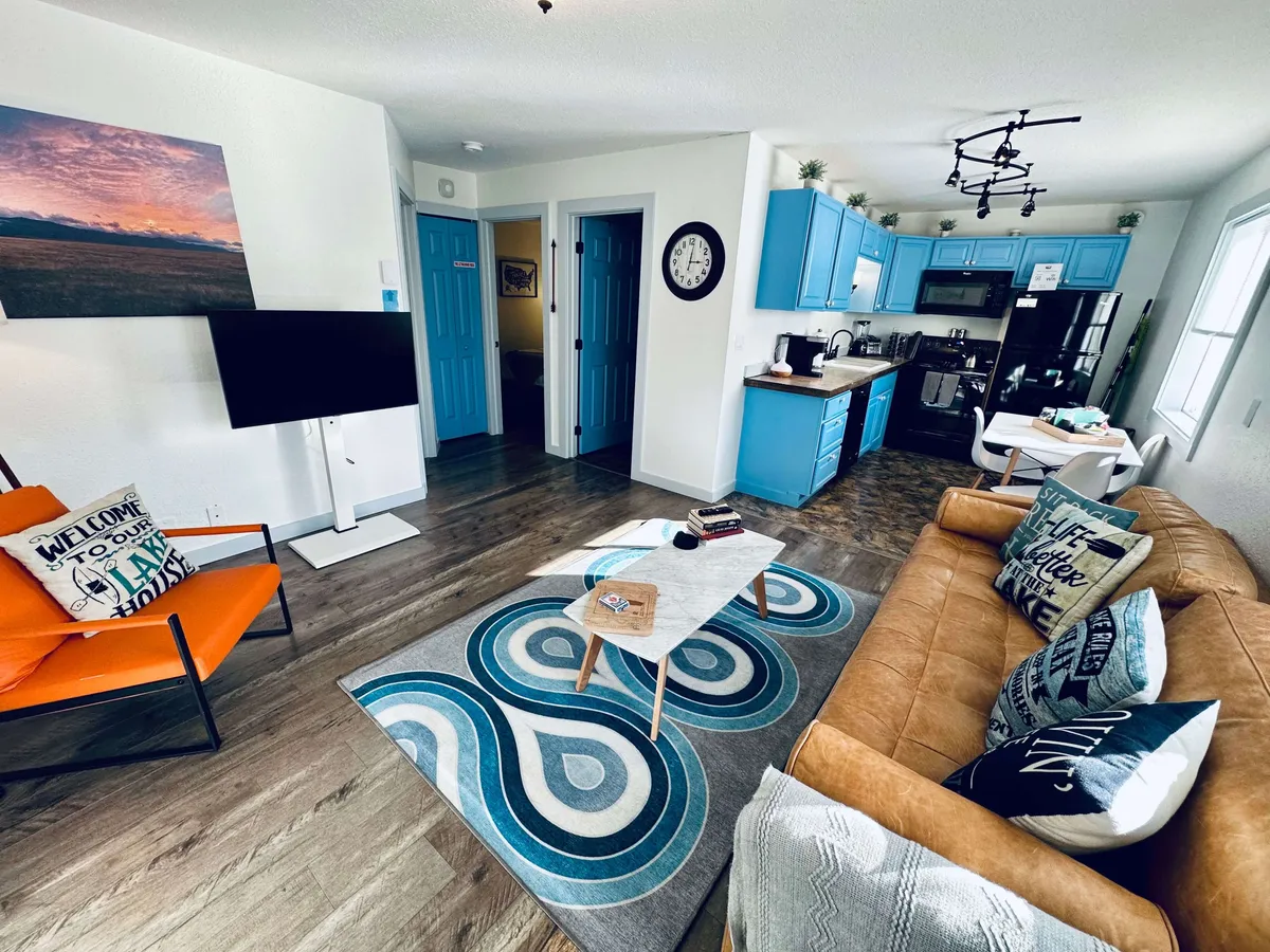 The image showcases a vibrant, inviting living space with a bold orange chair, striking blue kitchen cabinets, a cozy sofa adorned with patterned cushions, a coffee table topped with books, and a decorative rug. The scene hints at comfort, warmth, and relaxation, set in an open-concept layout perfect for gathering, unwinding, and enjoying time together.