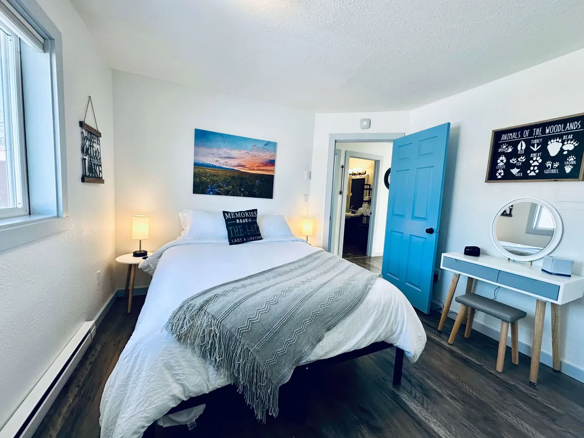 A serene bedroom features a bed with decorative pillows, a blue door, elegant wall art, and a vanity table with mirror. The space is bathed in natural light and exudes a calm, restorative vibe—perfect for relaxing or preparing for the day.