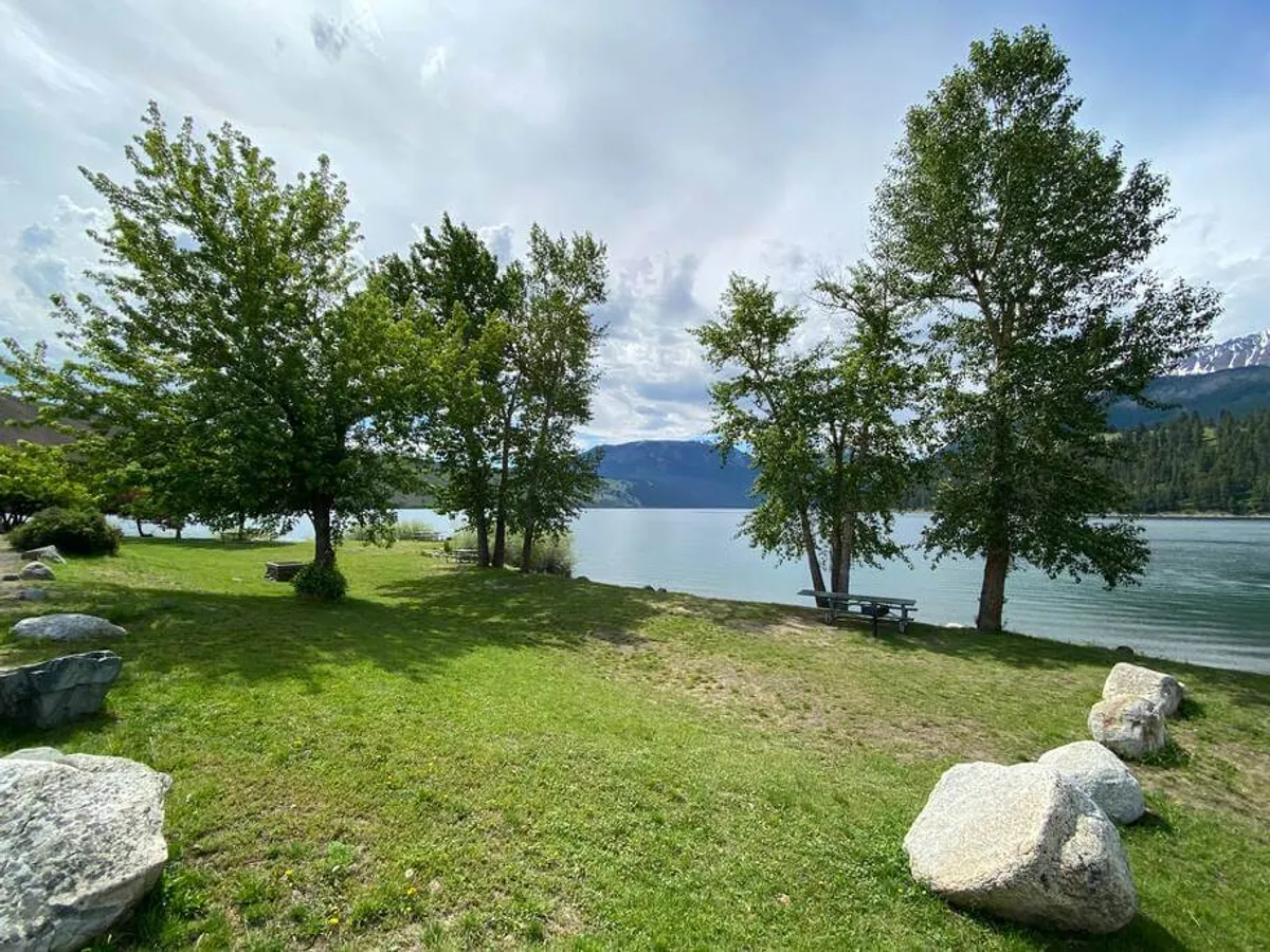 This image captures a tranquil lakeside scene with mature trees, rocks, and a designated picnic area. The water glimmers in the background, suggesting a peaceful space ideal for family picnics, fishing, or simply relaxing by the shore. The natural setting directly evokes the lakeside activities described in the section.