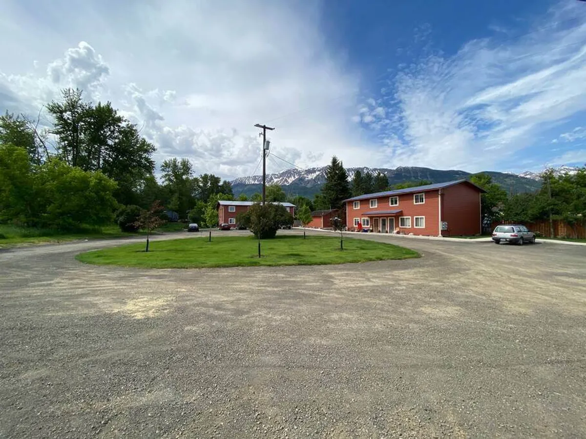 Red buildings set against a backdrop of green grass and majestic mountains, beneath a cloudy sky. The inviting open space encourages outdoor relaxation and serves as a launch point for nearby trails or park visits. Activities like picnicking or stargazing are suggested by the tranquil setting.
