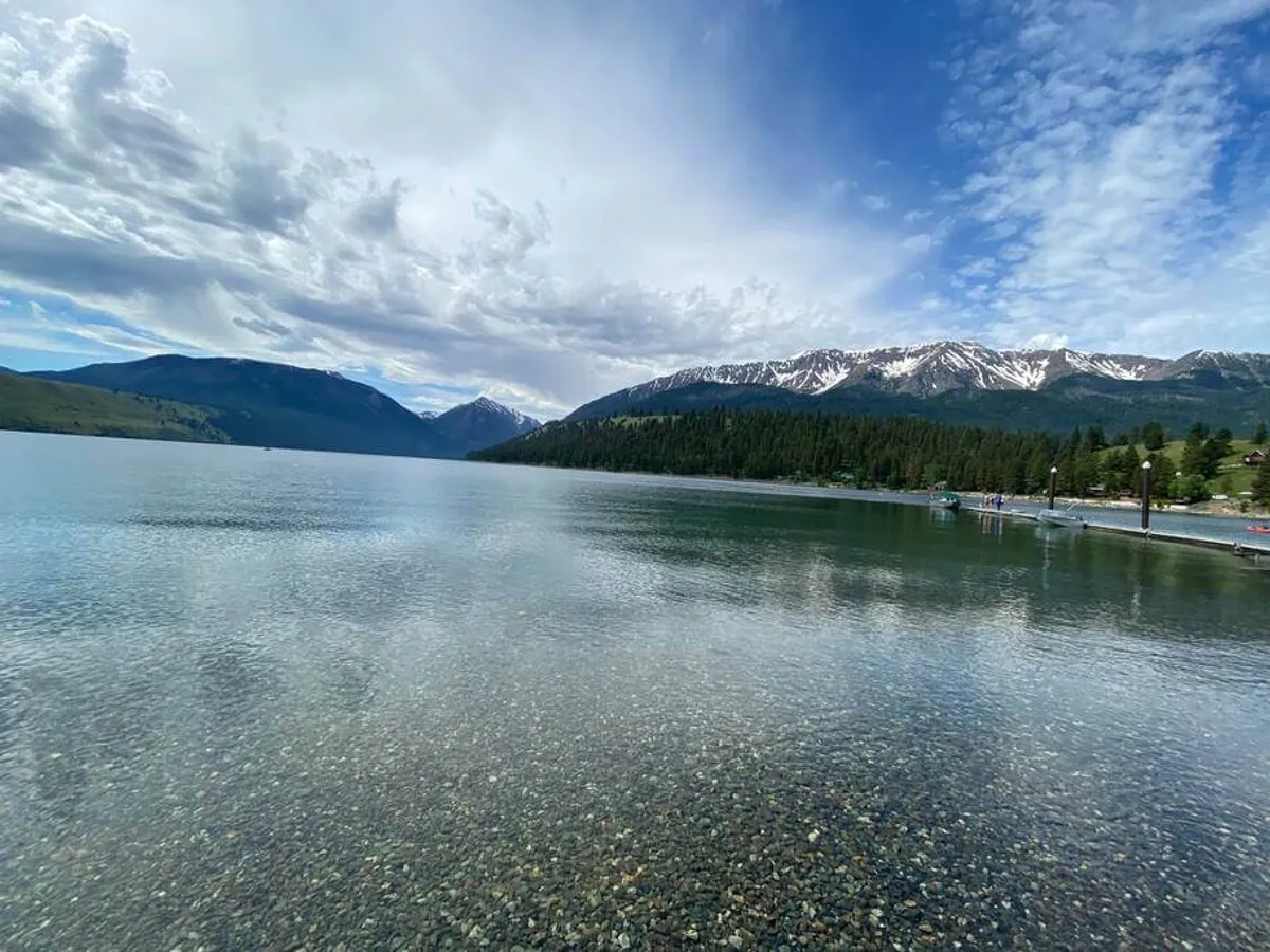 A breathtaking view across a calm lake ringed by lush green forest, with distant snow-capped mountains under a cloudy sky. The tranquil waters mirror the sky and peaks, evoking a sense of untouched wilderness and outdoor adventure. People can be seen enjoying boating and fishing along the shoreline, hinting at the recreational activities available.