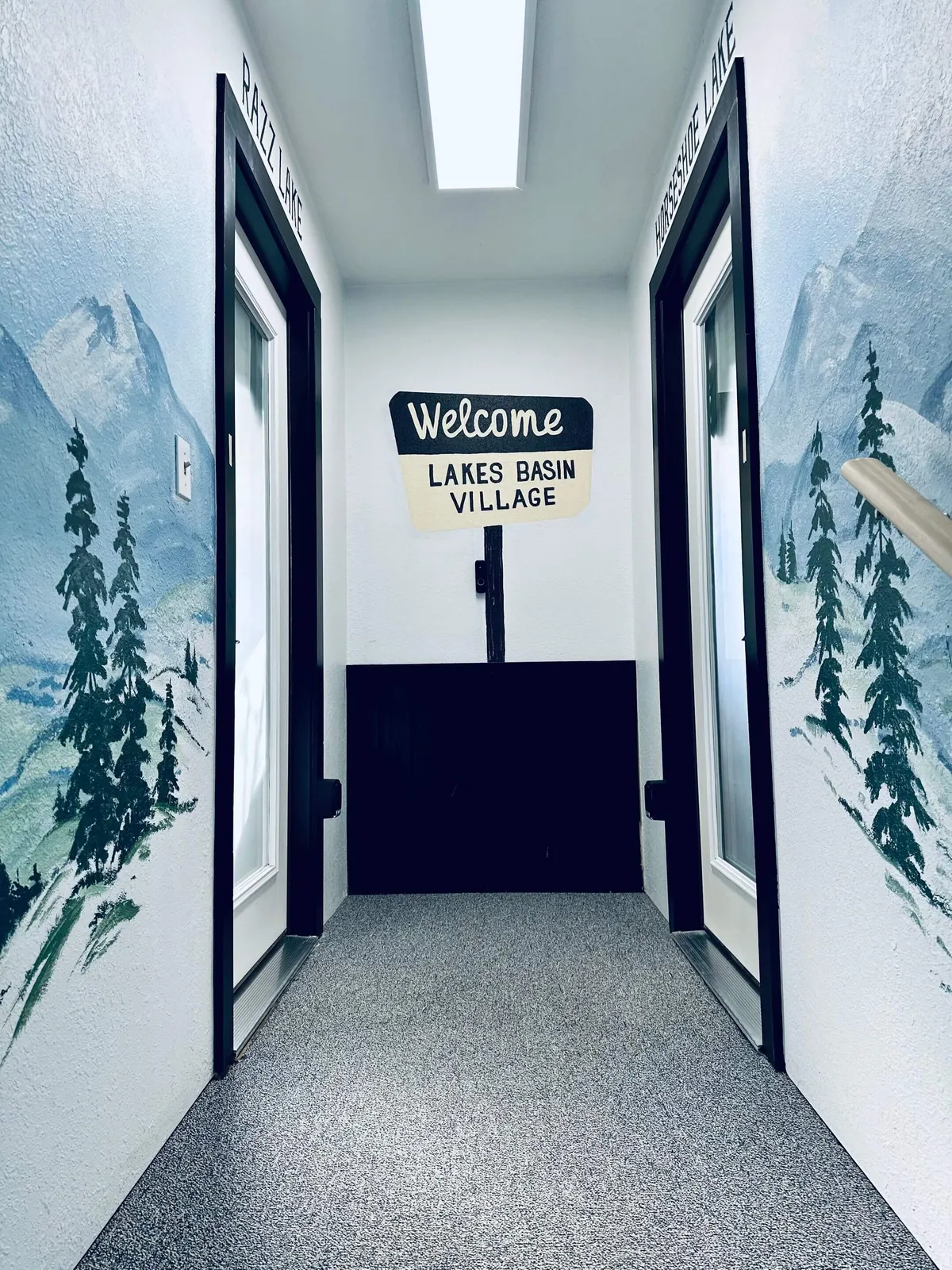 An inviting entryway area with a charming welcome sign, painted mountain scenery, and labeled doors provides a cozy transition space. It hints at the broader hospitality and communal spirit of the home, where guests gather information, plan activities, or simply settle in before heading to the kitchen or dining area.