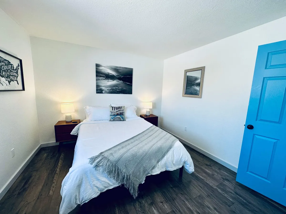 A tranquil bedroom scene featuring a plush bed adorned with crisp linens, a soft glow of natural light filtering through a window, a tasteful piece of artwork above the headboard, and a distinctive blue door lending character to the space. The nightstand is set for comfort, evoking a sense of calm and relaxation.